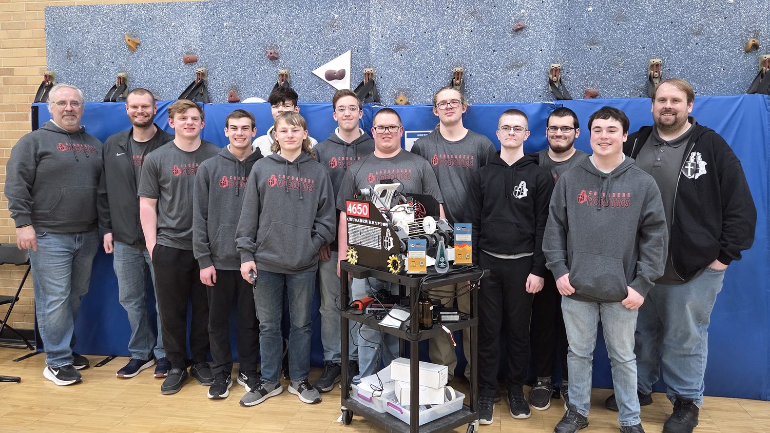 Team 4650 Kryptos students and their coaches in matching Crusader Robotics hoodies, gathered behind their robot cart at a competition